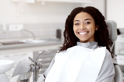 Smiling dental patient sitting in a chair wearing a bib, with dental equipment visible in a clean clinic setting