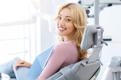 Smiling blonde woman reclining in a dental chair while wearing a protective bib in a bright treatment room.